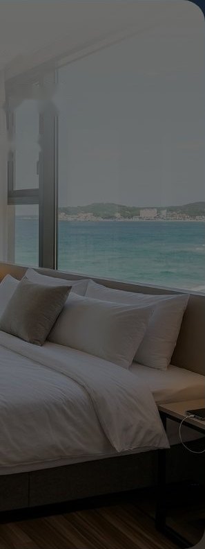 호텔 객실 — 흰 침구와 회색·흰 베개가 쌓인 침대, 협탁 위 전화기, 나무 바닥, 통창 너머 청록빛 바다와 먼 해안 마을