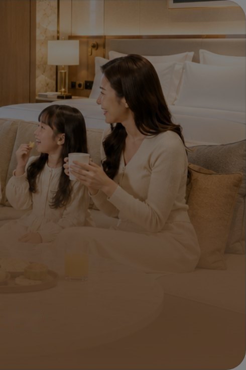 호텔 객실 침대에 앉아 간식을 먹는 딸과 흰 머그컵을 든 엄마
