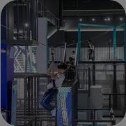실내 키즈 테마파크 — 안전 하네스를 착용한 아이가 청록색 철제 프레임의 어두운 실내 홀에서 로프 코스에 매달려 있고, 위쪽 플랫폼에 다른 사람이 서 있다