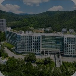 호텔 어라운드 평창 — 푸른 산림에 둘러싸인 길고 낮은 흰색 유리 호텔 항공 뷰, 왼쪽 고층 아파트, 흰 구름 낀 파란 하늘