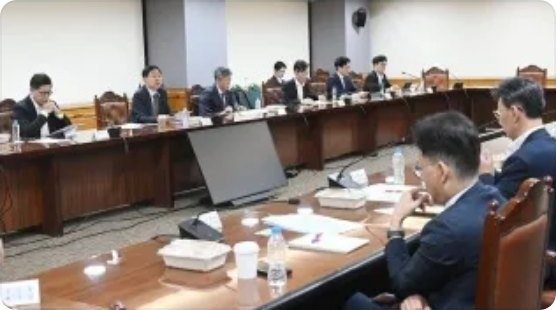 정장 차림 남성들이 마이크가 놓인 긴 원목 회의 탁자에 둘러앉아 회의하는 국회 회의장 내부