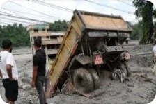 진흙과 화산재에 바퀴까지 파묻힌 노란 덤프트럭과 이를 지켜보는 두 남성