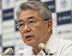 記者会見でマイクの前に座る白髪・眼鏡の年配男性（白いシャツ）