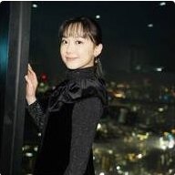 夜景を背景に黒いフリル付きの衣装で手を上げて微笑む若い女性タレントのポートレート